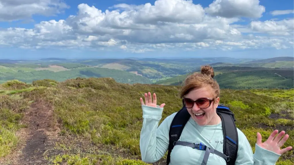 Ciara from the Earth's Edge team, wearing a backpack, out hiking with a view of mountains on a cloudy but sunny day.