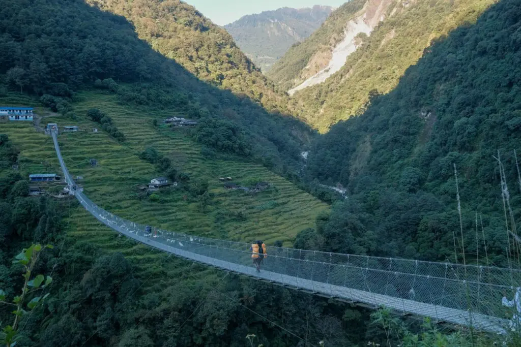 A porter carrying duffel bags crossing a suspension bridge over the lush Annapurna Sanctuary, Nepal