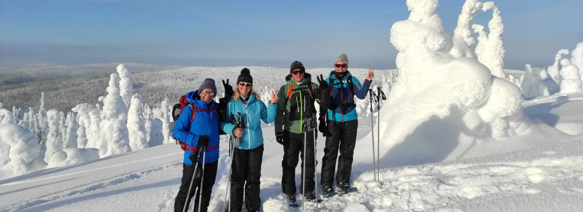 Four people snowshoeing in Lapland, Finland, posing in winter gear among snow-covered trees in the Arctic landscape under a clear blue sky.