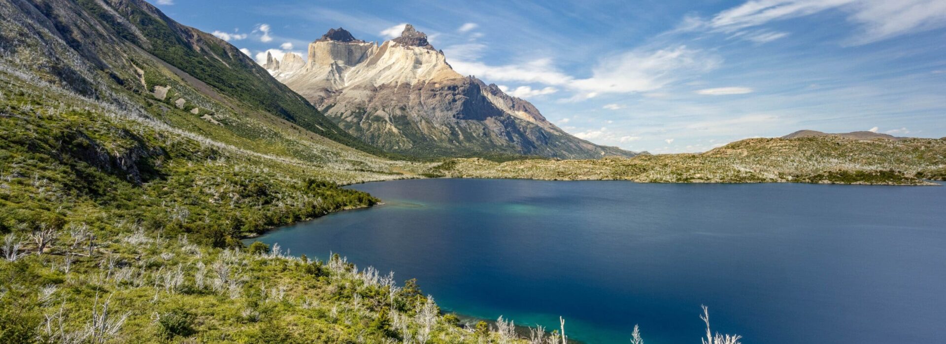 Panoramic view across a deep blue Patagonian lake toward the Paine massif and Cuernos del Paine peaks under a bright summer sky.