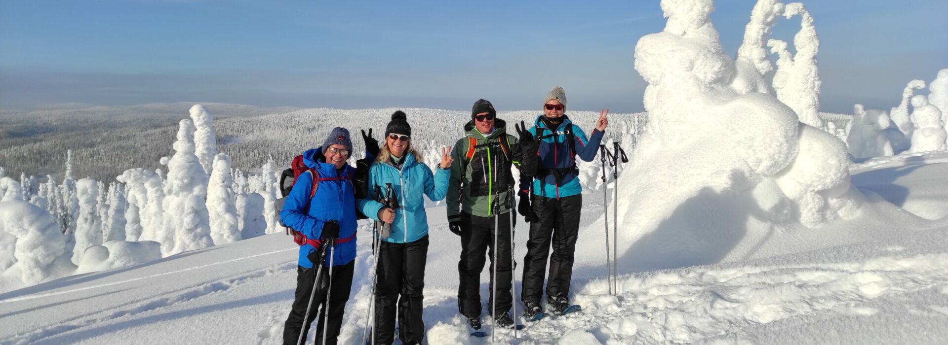 Four people snowshoeing in Lapland, Finland, posing in winter gear among snow-covered trees in the Arctic landscape under a clear blue sky.